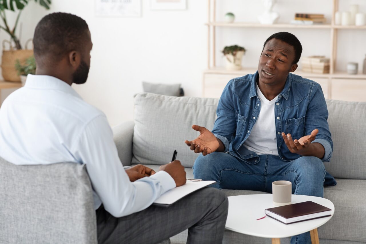 Stressed black man explaining his problems to psychologist at individual therapy session at office, copy space
